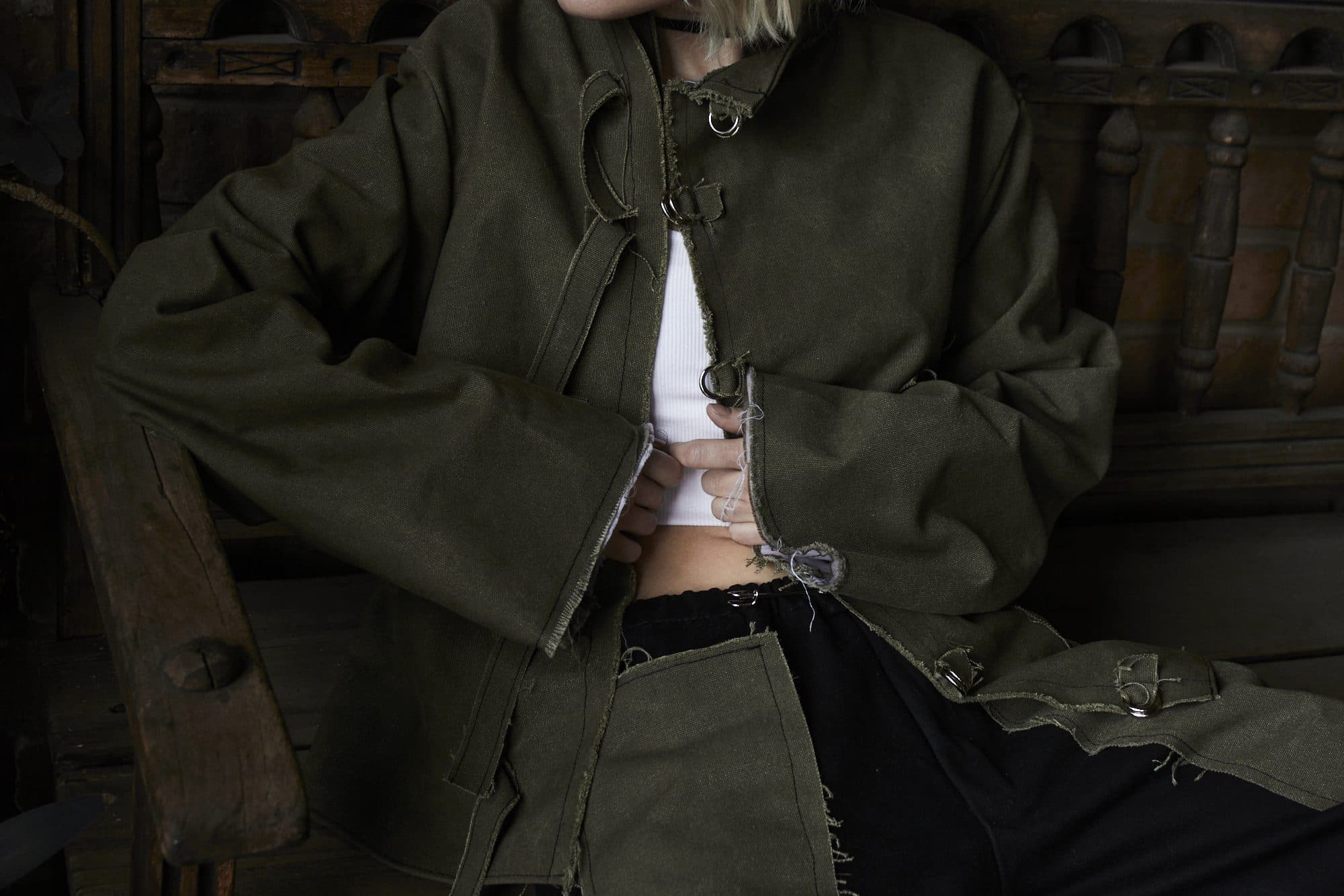 Close-up of the AKIN jacket: raw edges, rings and loops, worn over a white top