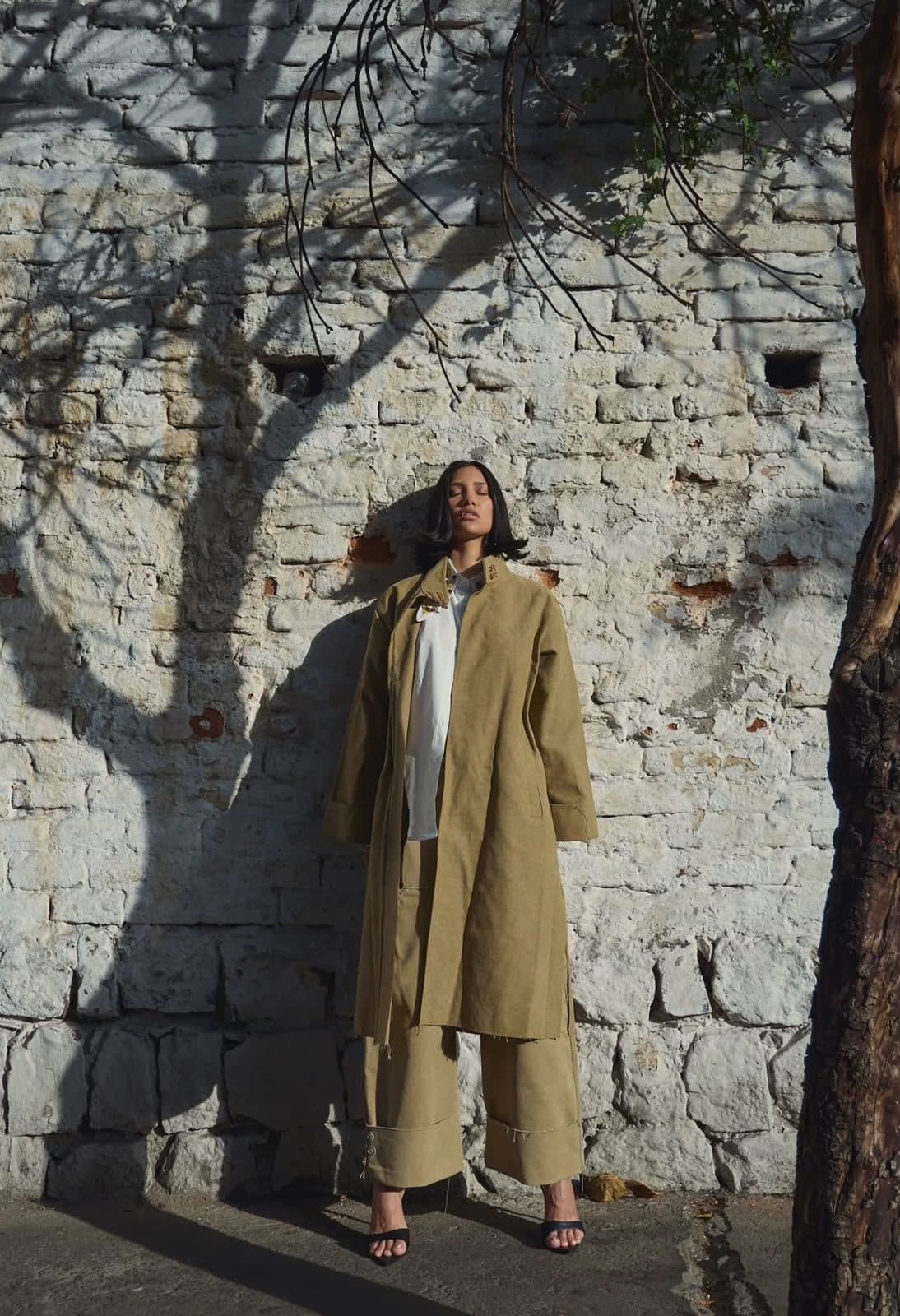 The FRAME coat and trousers worn against a whitewashed brick wall, under the shadow of a tree