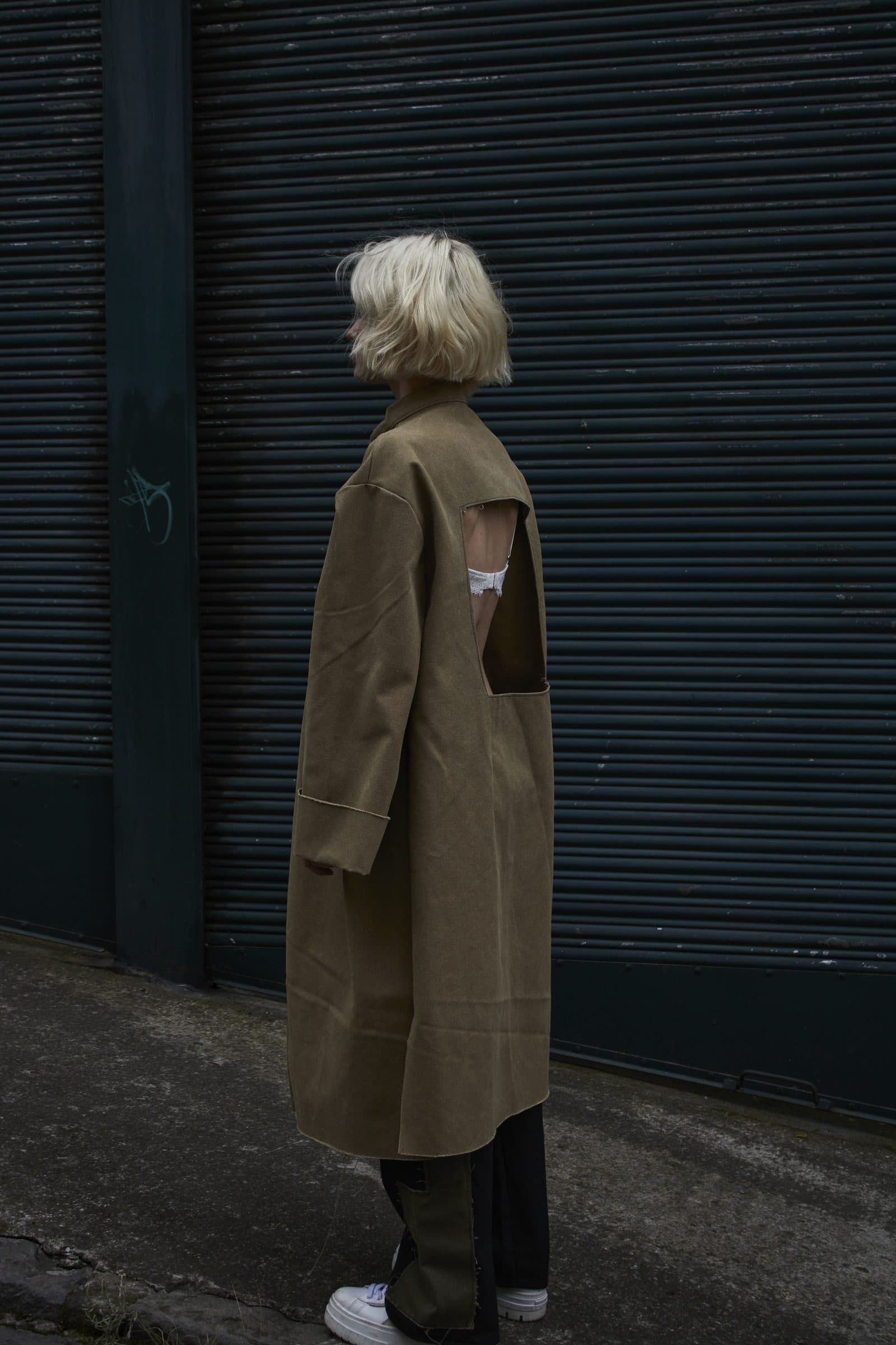 FRAME coat from behind, with the open back and a white label, against a dark shutter